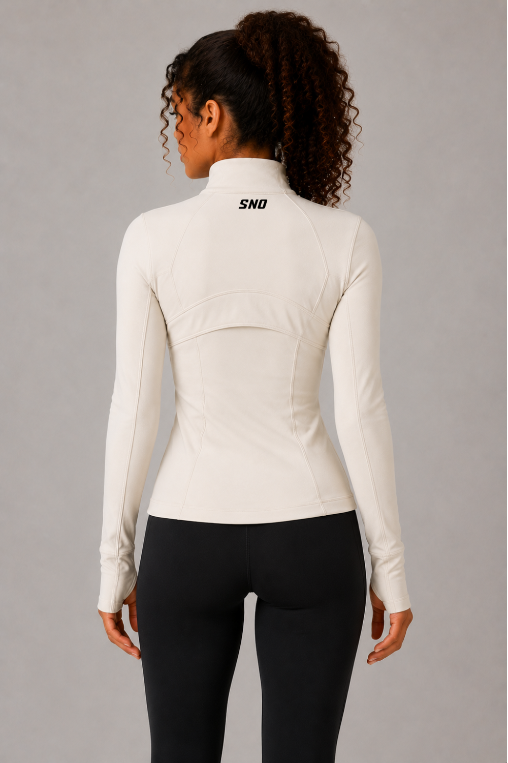 A woman shown from the back wearing a fitted long-sleeve athletic top in off-white with a small black 'SND' logo on the upper back, paired with black leggings, against a plain gray studio background.
