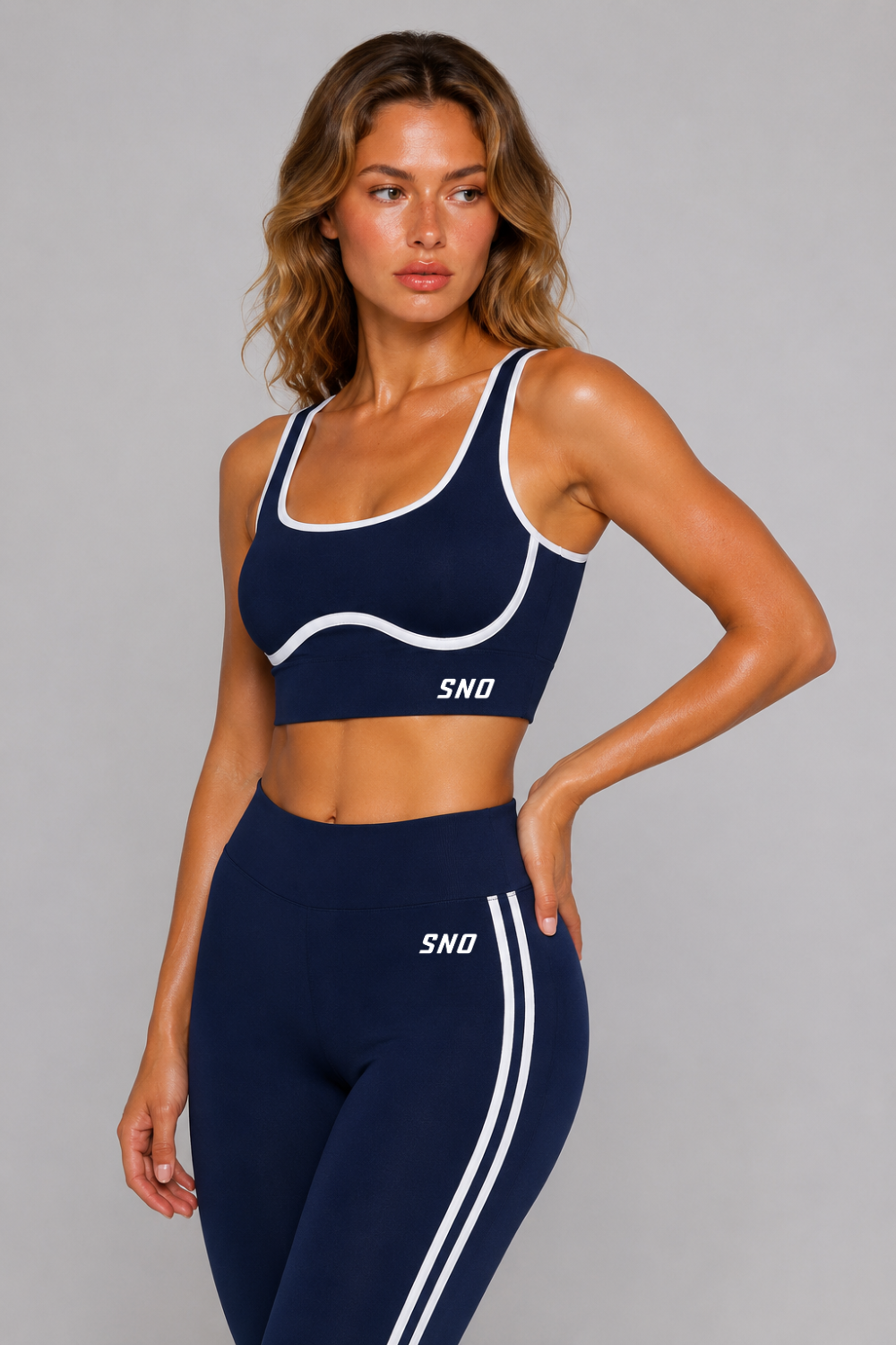 Woman wearing a navy sports bra and leggings set with 'SNO' branding on a gray background