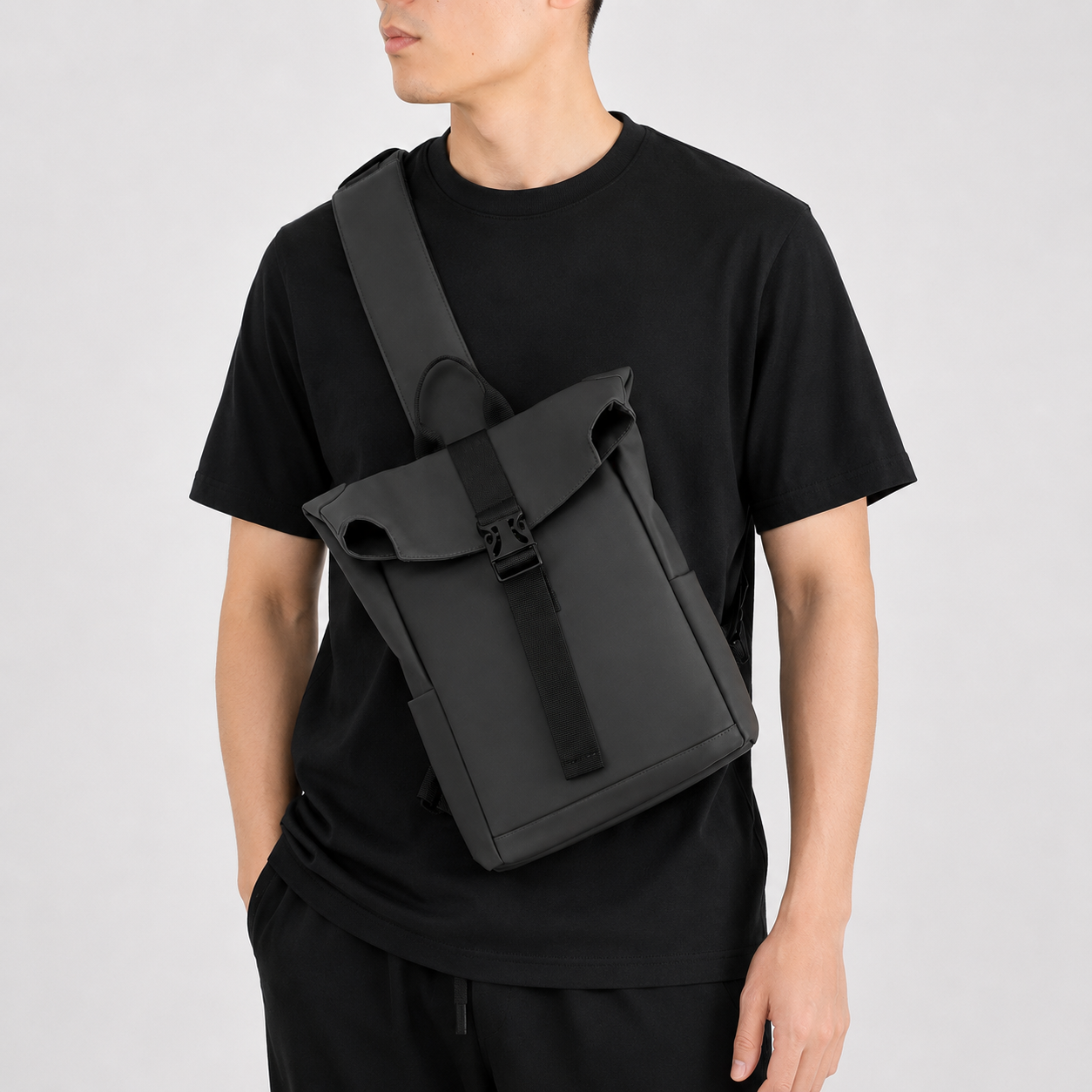 A man wearing a black t-shirt and black pants models a black roll-top sling backpack with a wide adjustable strap, set against a plain light gray background.