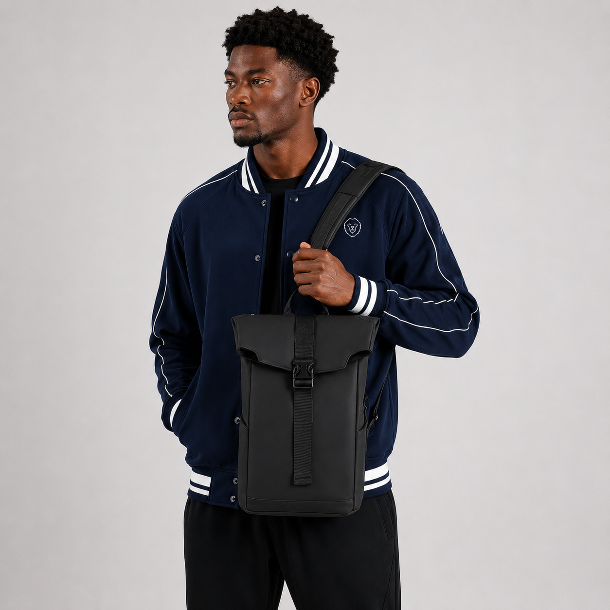 A man wearing a navy blue varsity jacket with white stripes and black pants, carrying a black roll-top backpack on his shoulder against a light gray studio background.